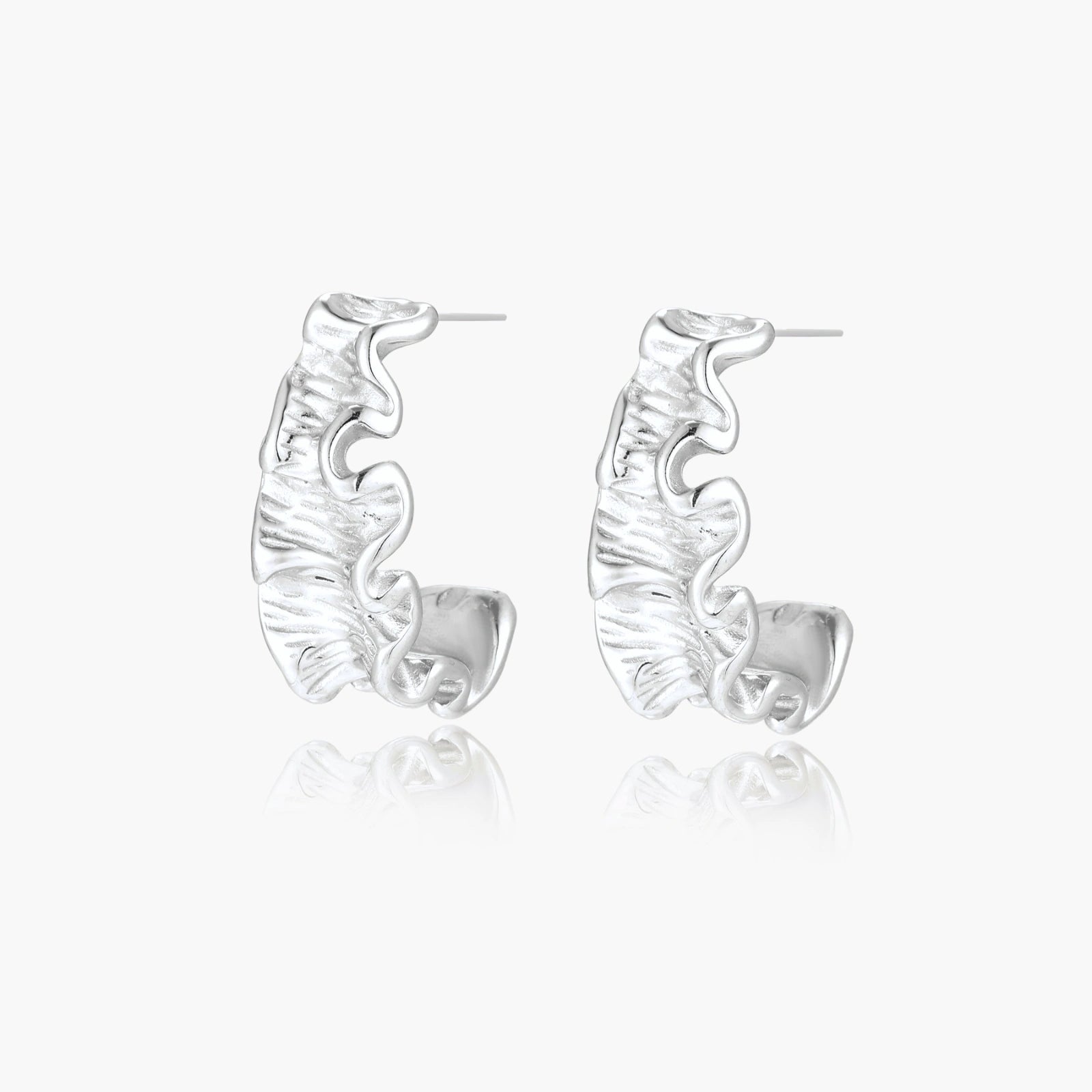 Textured white gold-plated hoop earrings with flowing wave pattern against cream background, showing dimensional rippled surface.