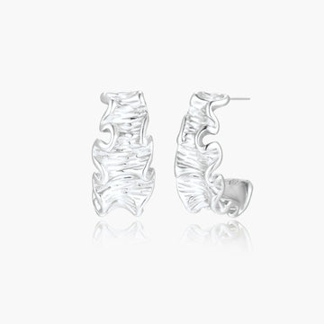 Textured white gold-plated hoop earrings with flowing wave pattern against cream background, showing dimensional rippled surface.
